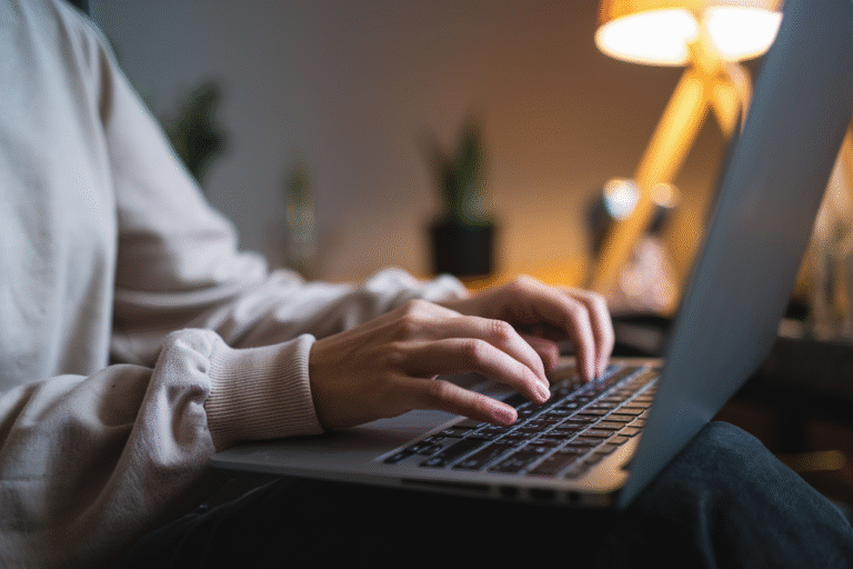 A close-up view of hands typing on a laptop keyboard. The person is wearing a light beige long-sleeve sweatshirt.